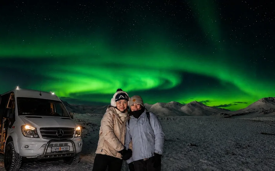 Ein 4x4-Minibus unter einem grünen Polarlicht mit zwei Reisenden daneben